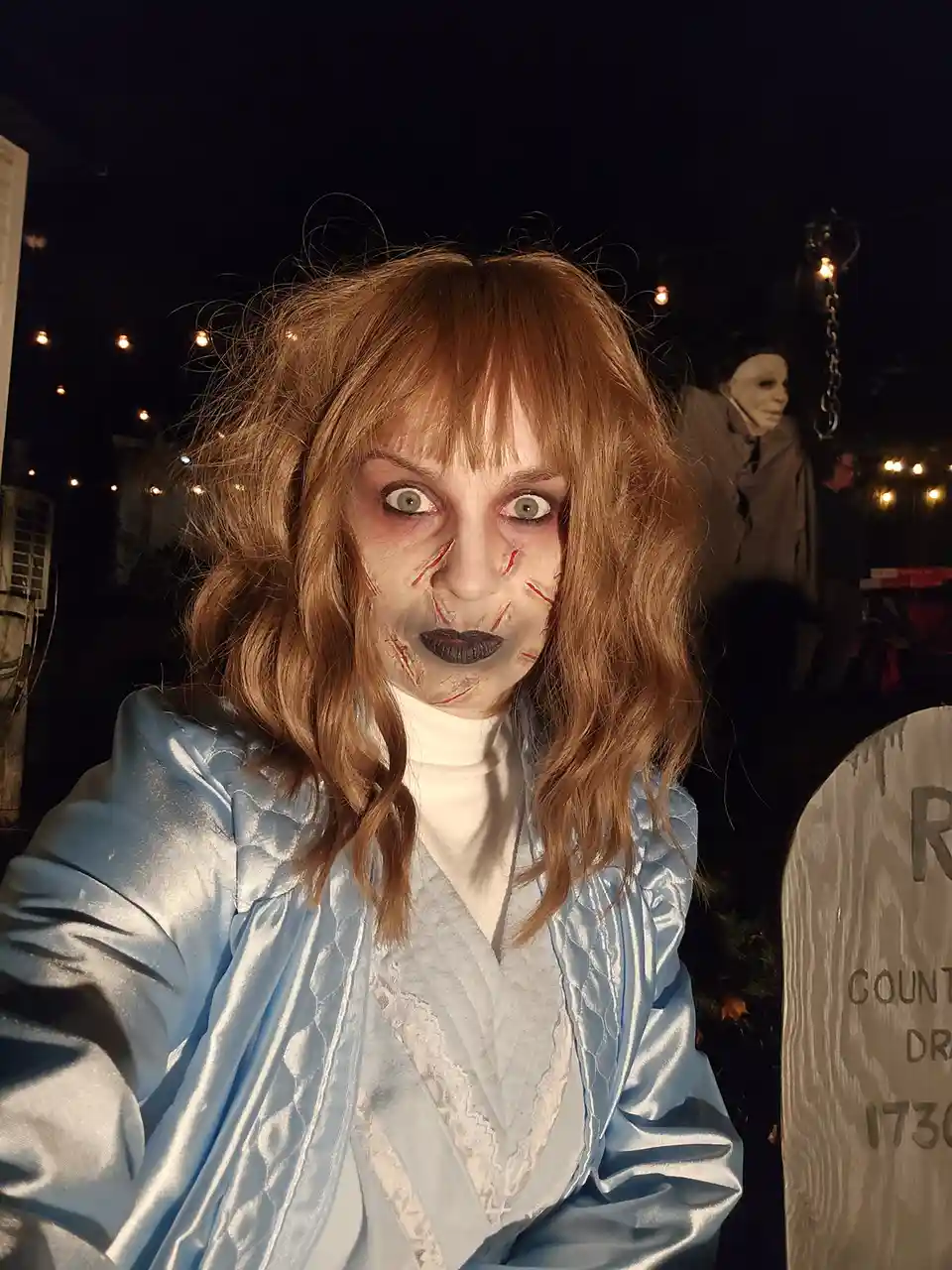 A person in a zombie costume with pale makeup, red stitched wounds, and wide eyes takes a selfie at night. They wear a light…