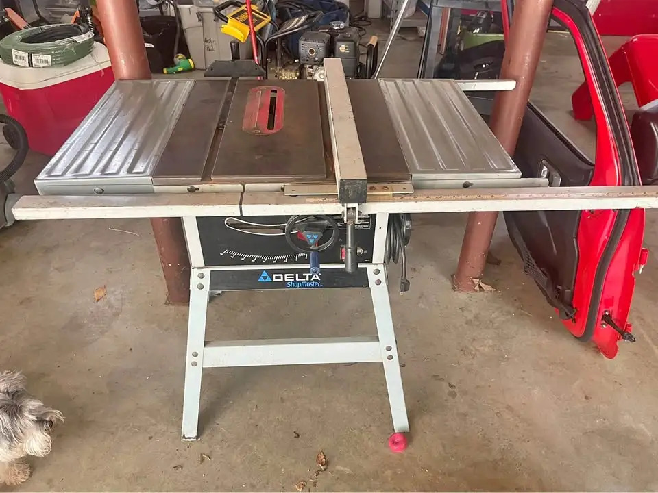A Delta table saw sits in a cluttered garage, with a long wooden plank resting across its surface. To the right, a red car…