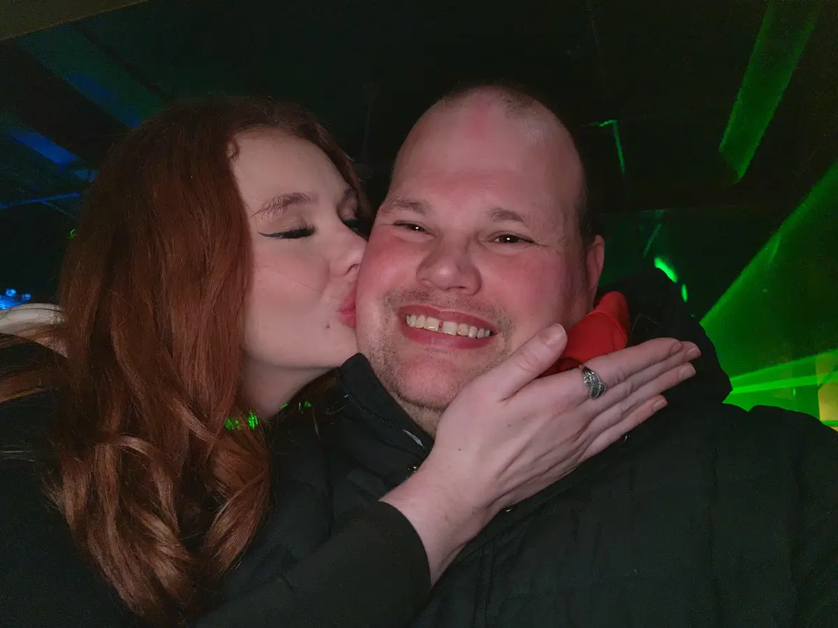 A woman with long red hair kisses a smiling bald man on the cheek while holding his face; he wears a black jacket and has a…