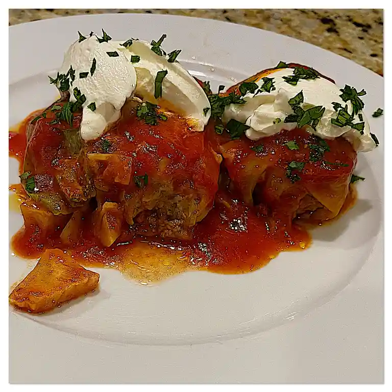 A close-up photo shows two large, messy, sauce-covered stuffed cabbage rolls on a white plate, topped with dollops of sour…