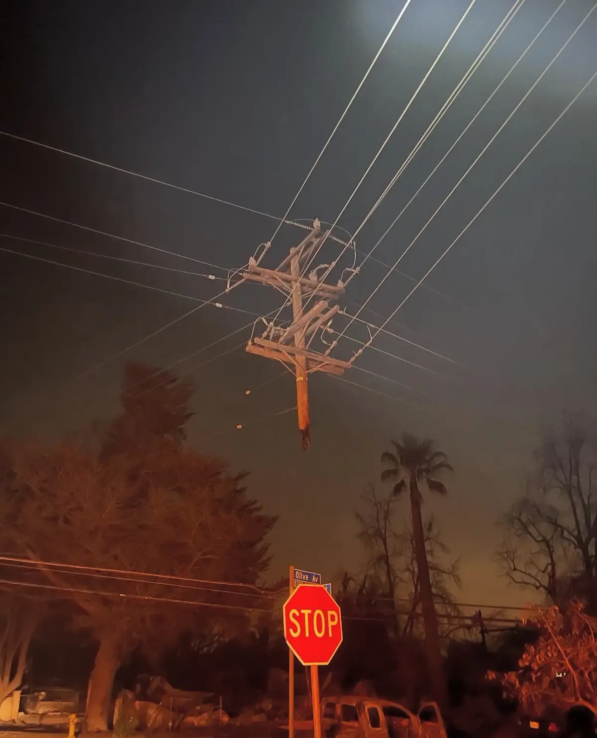 A nighttime photo shows a wooden utility pole hanging precariously by wires against a smoky, orange-hued sky, with a red…