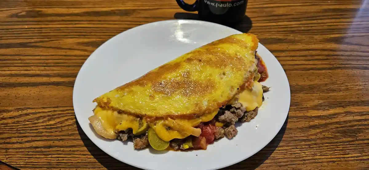 A close-up photo shows a folded, golden-brown omelette on a white plate, revealing melted cheese, ground meat, diced…