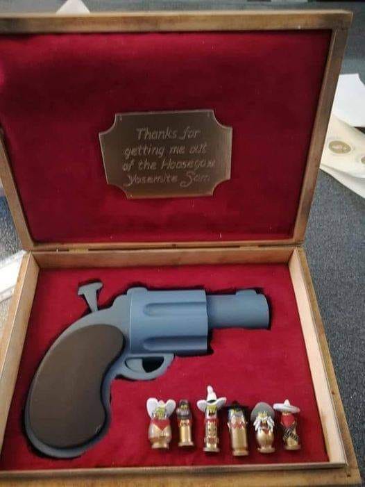 An open wooden presentation case with red velvet lining contains a large, gray toy revolver and five small, decorative…