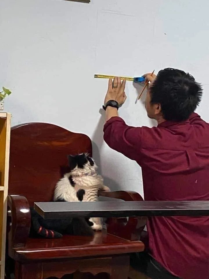 A person in a maroon shirt is measuring a white wall with a yellow tape measure while holding a pencil, with a…