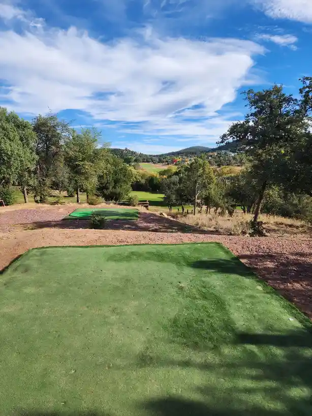 A scenic golf course view with two green practice mats on reddish-brown gravel, surrounded by trees under a blue sky with…