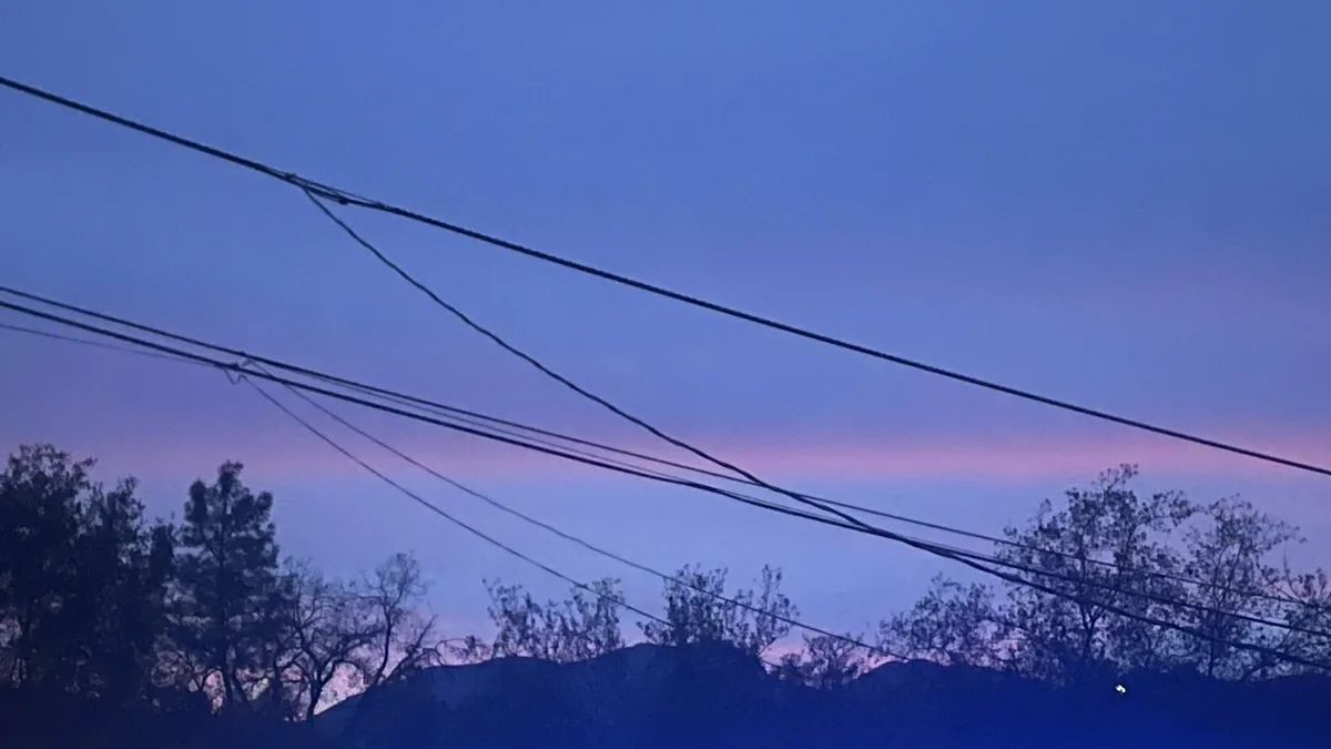 A photo of a twilight sky with purple and blue hues, silhouetted trees, and multiple power lines crossing the frame…