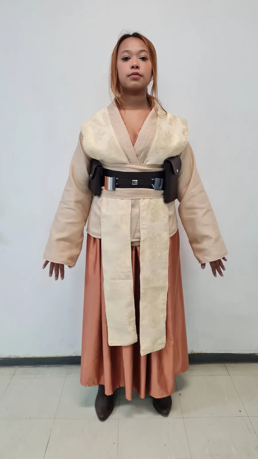 A person stands facing the camera in a full Star Wars Jedi robe costume with a utility belt, looking blankly at the viewer…