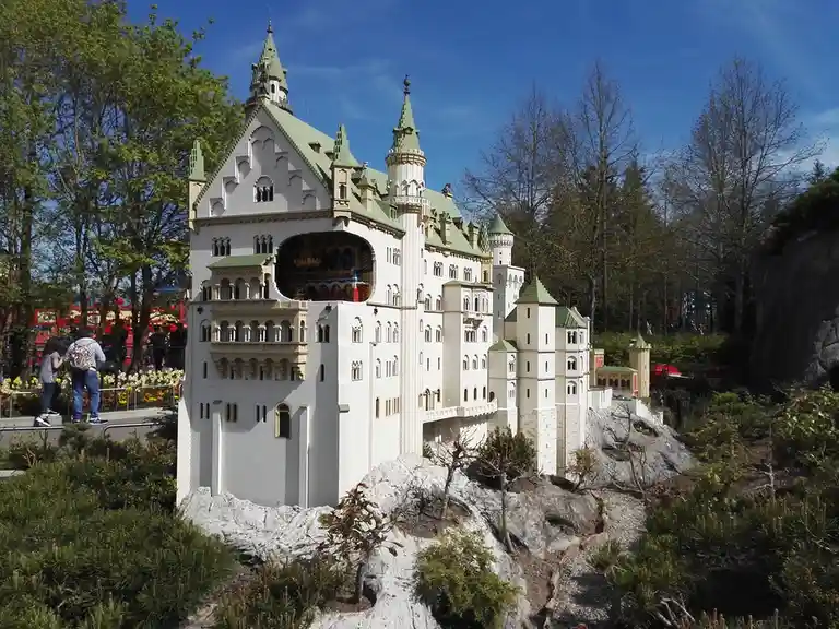 a large, detailed miniature castle with white walls and green roofs, built from lego bricks, sits on a rocky hillside…
