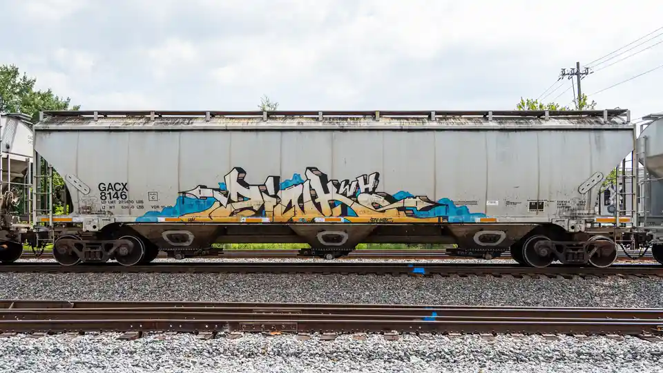 A weathered white railroad hopper car sits on tracks under a cloudy sky, featuring large, stylized graffiti in beige, black…