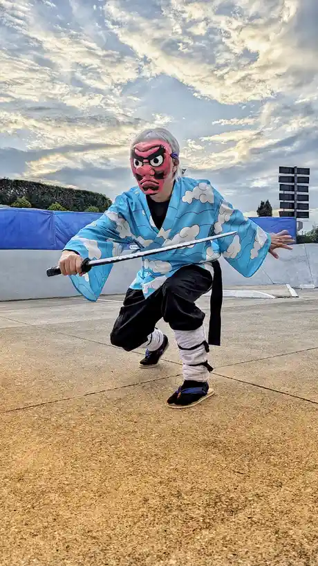 A person in a blue cloud-patterned kimono and red demon mask strikes a dynamic martial arts pose outdoors, holding a sword…