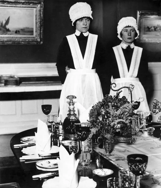 Two women in vintage maid uniforms stand behind an elegantly set dining table with glassware, plates, and a centerpiece. The…