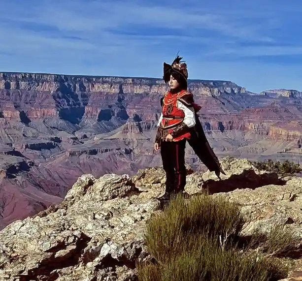 A person in ornate red and black fantasy armor, complete with a feathered hat, stands on a rocky overlook of the Grand…