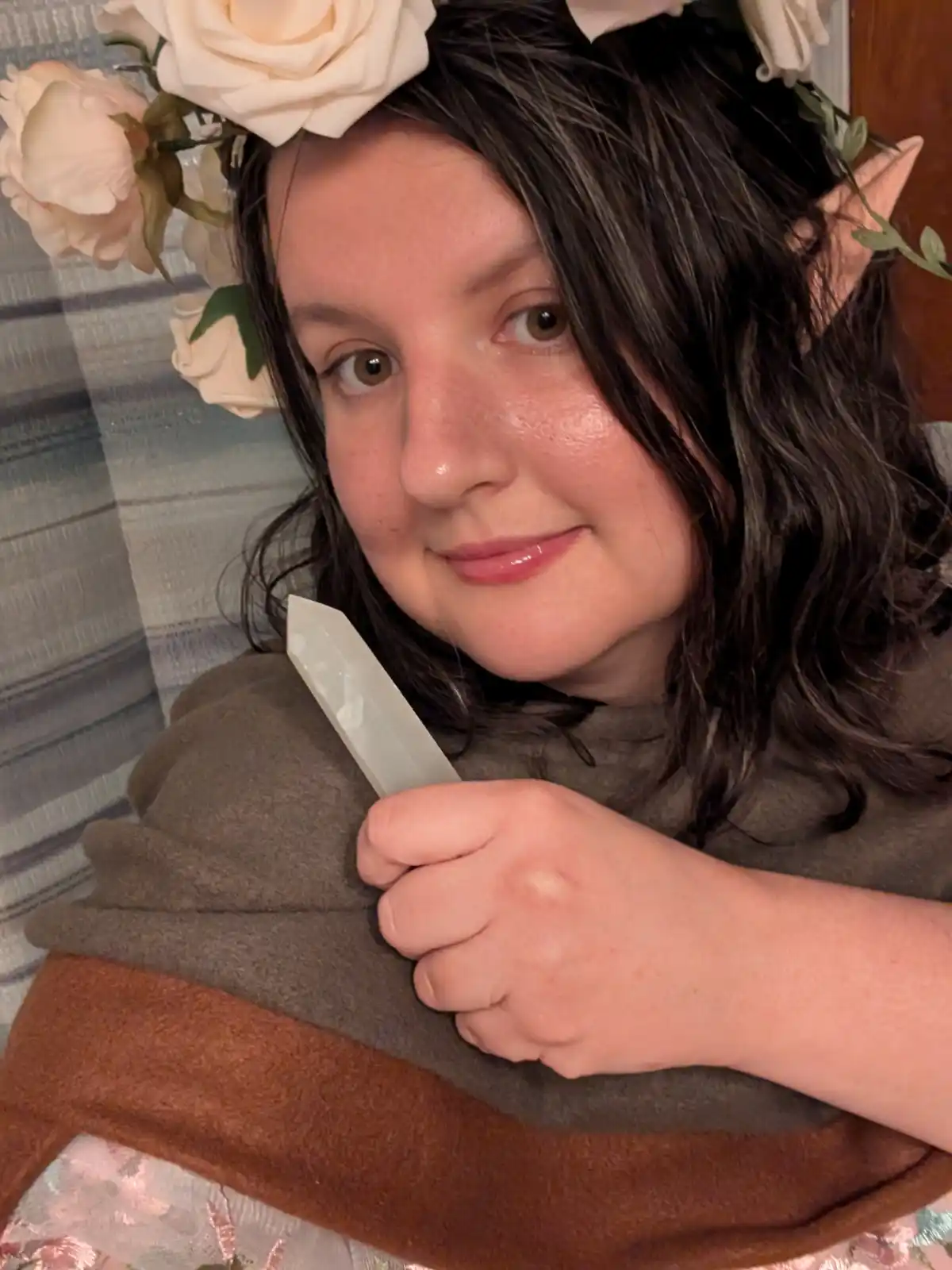 A person with dark hair, wearing a floral headpiece and pointed elf ears, holds a translucent crystal wand while looking at…