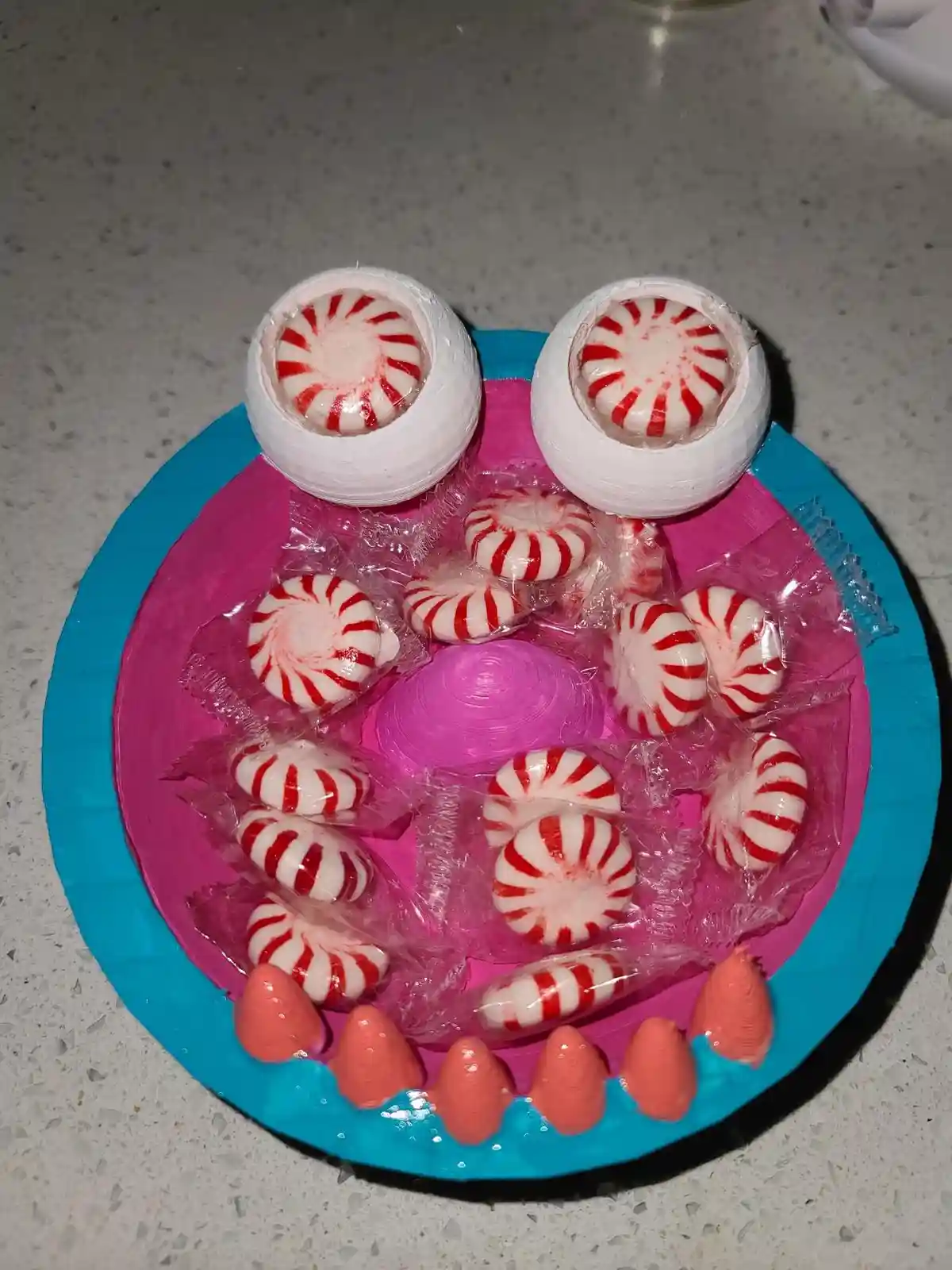 A handmade craft resembling a monster's face on a blue paper plate, featuring two large white spheres with peppermint…