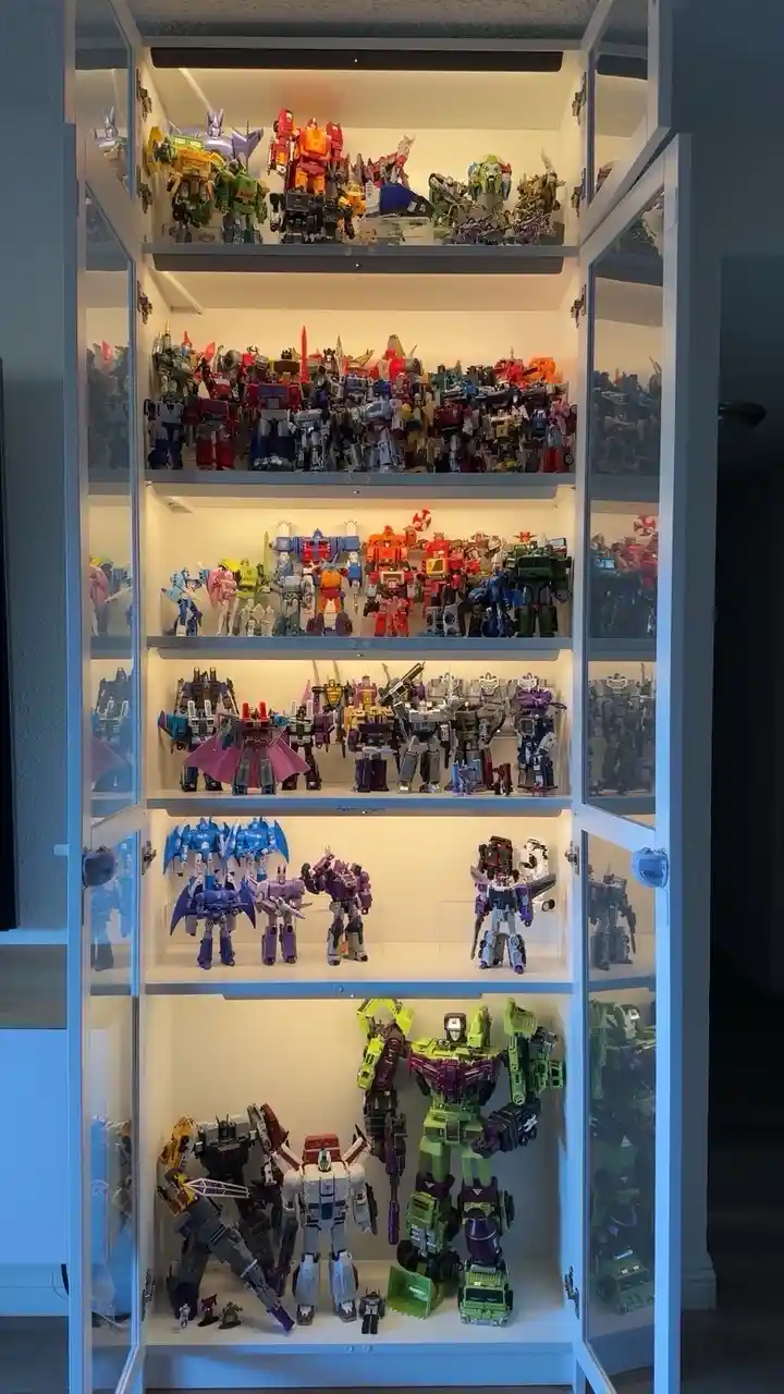 A tall, white, glass-fronted cabinet with multiple shelves is filled with hundreds of colorful Transformer action figures…