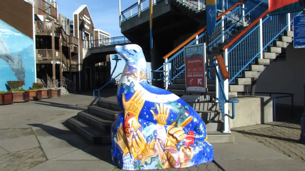 A large, colorful, painted otter statue sits on a sidewalk near stairs and buildings, featuring various animals and hands on…