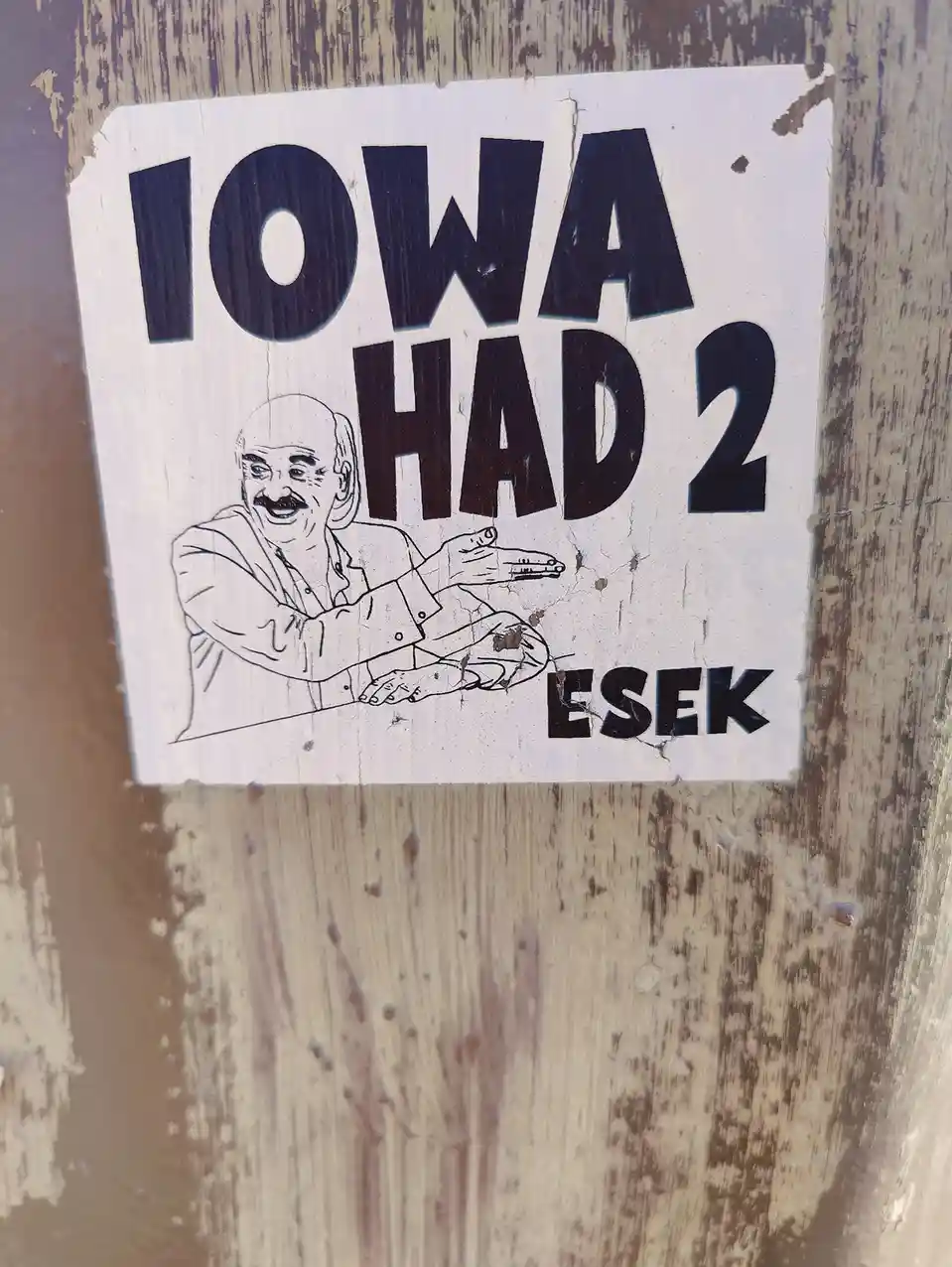 A weathered white sticker on a wooden surface features bold black text reading 'IOWA HAD 2 ESEK' and a line drawing of a…