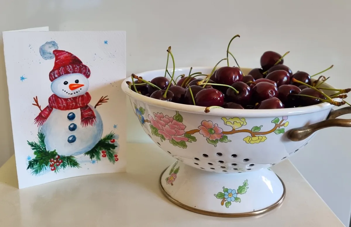 A festive snowman greeting card stands beside a white ceramic colander filled with fresh, dark red cherries with stems. The…