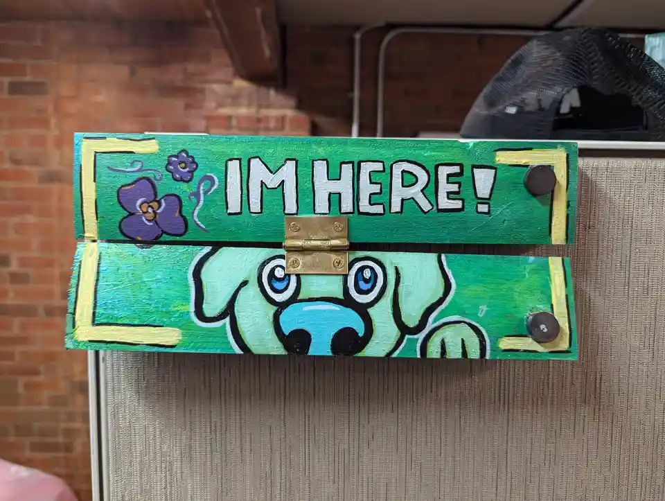 A handmade green wooden sign with a brass hinge, painted with a cartoon dog's face peeking out from below and purple flowers…