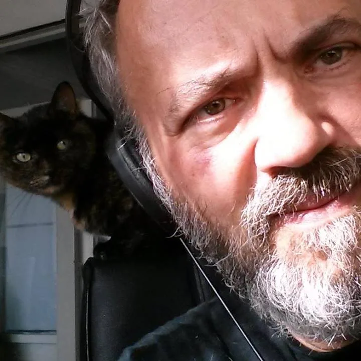 A close-up selfie of a man with a gray beard and headphones, looking directly at the camera with a stern, disappointed…