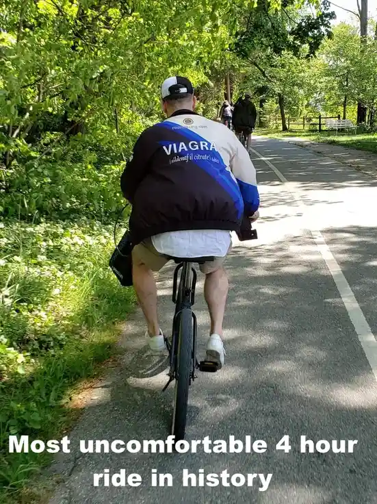 A man is seen from behind riding a bicycle on a paved path through a sunny, tree-lined park. He wears a black, white, and…