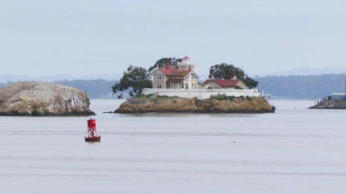 A scenic photo shows a lighthouse complex on a rocky island in the water, with a red buoy marked '18' floating nearby. The…