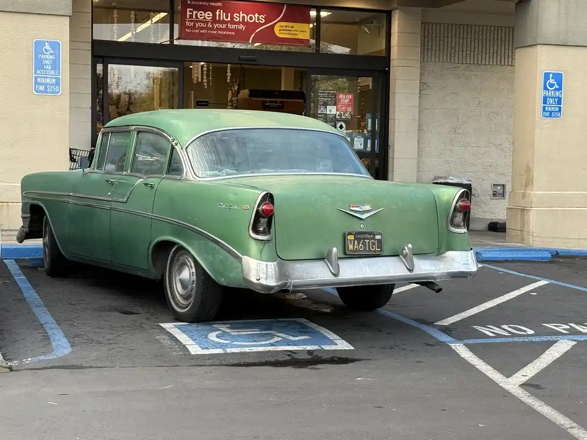 A weathered green 1950s Chevrolet Bel Air is parked in a designated handicapped parking space marked with a blue wheelchair…