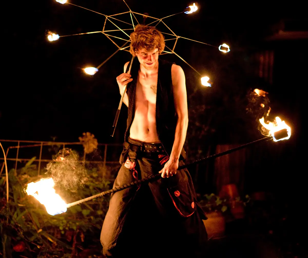 A shirtless man with curly hair, wearing a black vest and pants, performs fire dancing at night. He holds a long pole with…