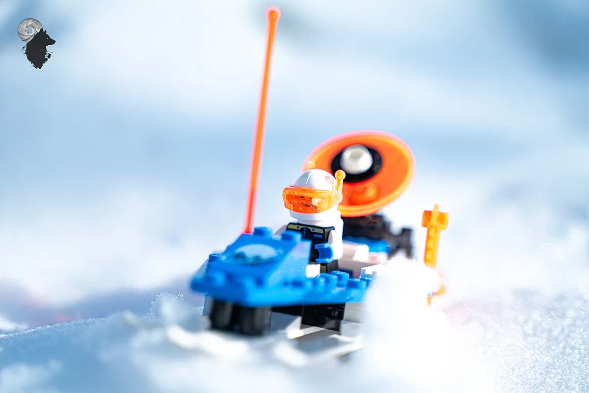 A close-up photo of a LEGO minifigure wearing an orange helmet and goggles, seated on a blue LEGO snowboard with an orange…