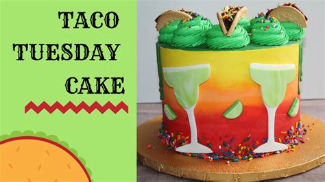 A colorful cake decorated with green frosting, two margarita glasses, lime wedges, and small taco-shaped toppers, sits on a…