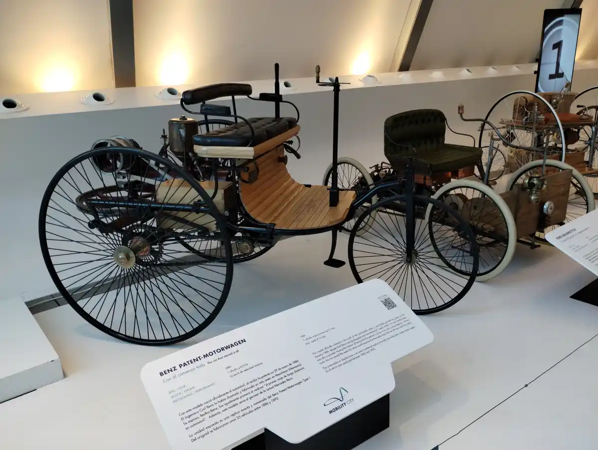 A museum display features a replica of the Benz Patent-Motorwagen, the first automobile, with large spoked wheels, a wooden…