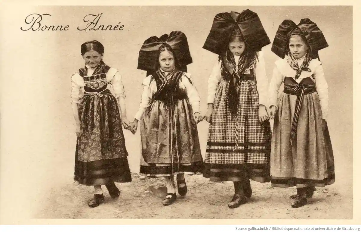 A vintage sepia-toned photograph shows four young girls in traditional folk costumes holding hands. Three of the girls wear…
