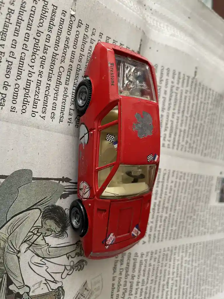 A red toy race car with visible wear and tear, including a large gray patch on its side, is positioned on a newspaper. The…