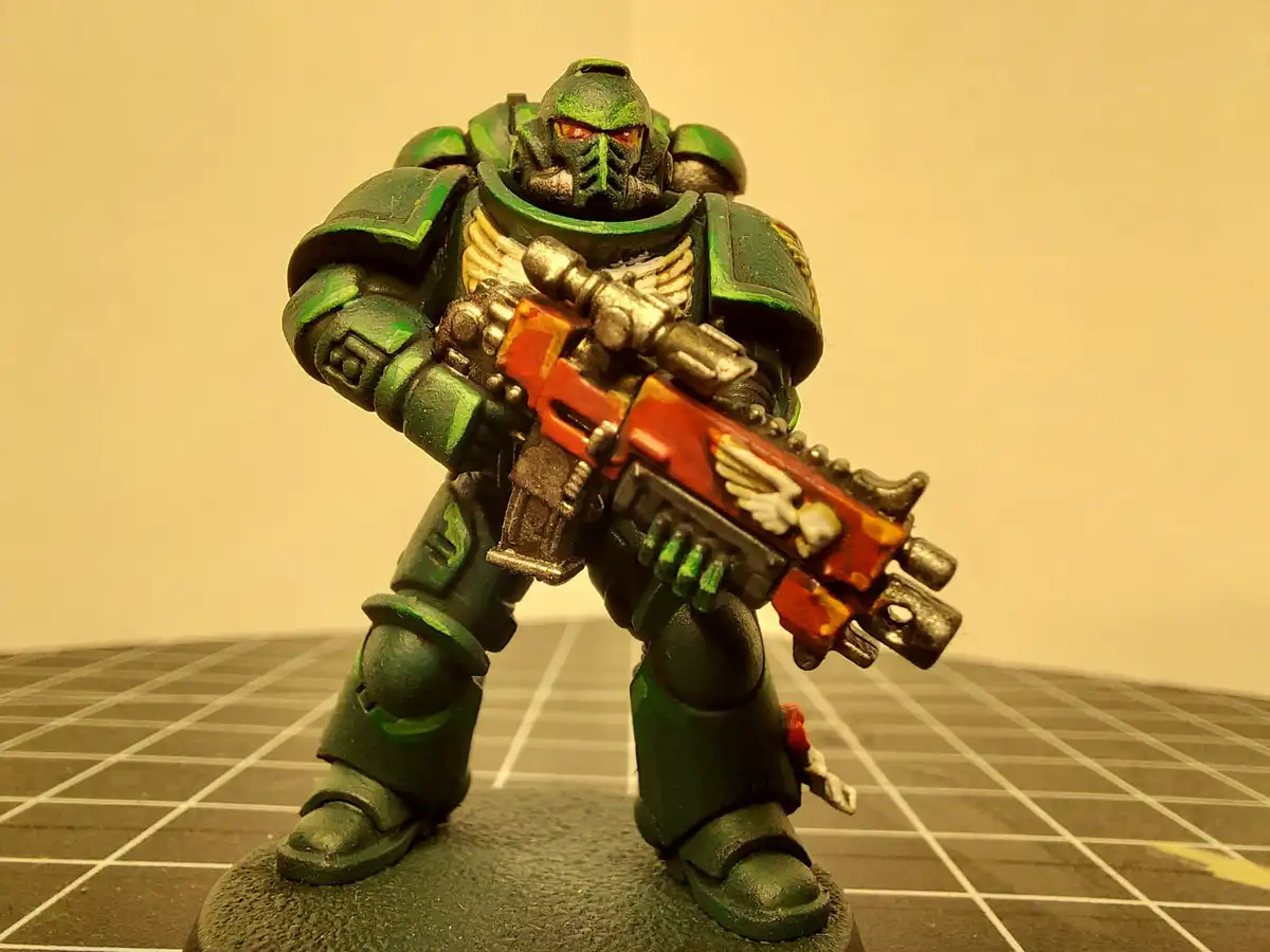 A painted miniature of a green-armored Space Marine from Warhammer 40,000 stands on a grid mat, holding a red and orange…