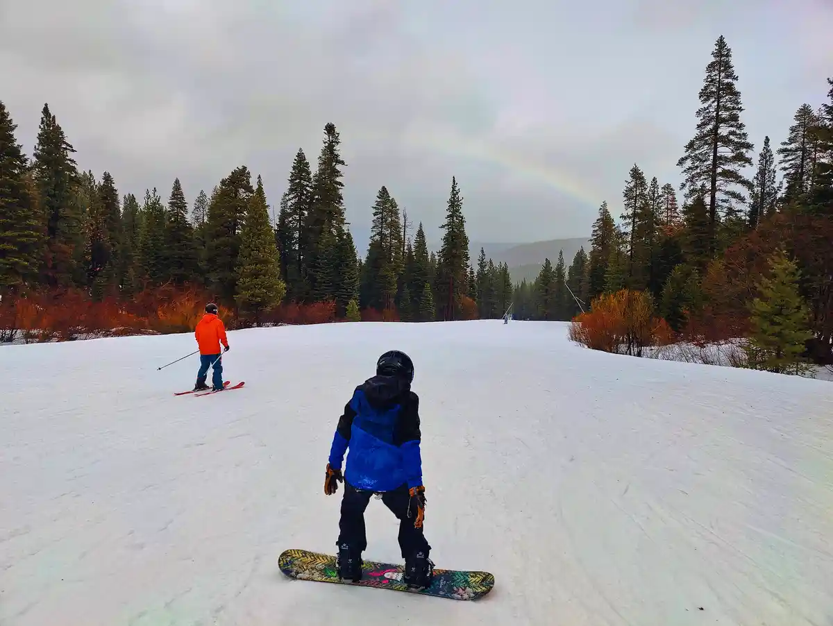 Two people are on a snowy slope, one on skis and one on a snowboard, both facing away from the camera toward a faint rainbow…