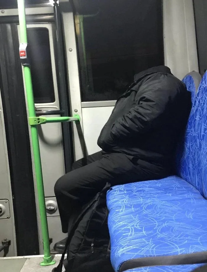 A person in a black jacket and pants is asleep on a blue patterned bus seat, their head down and body slumped. A green pole…
