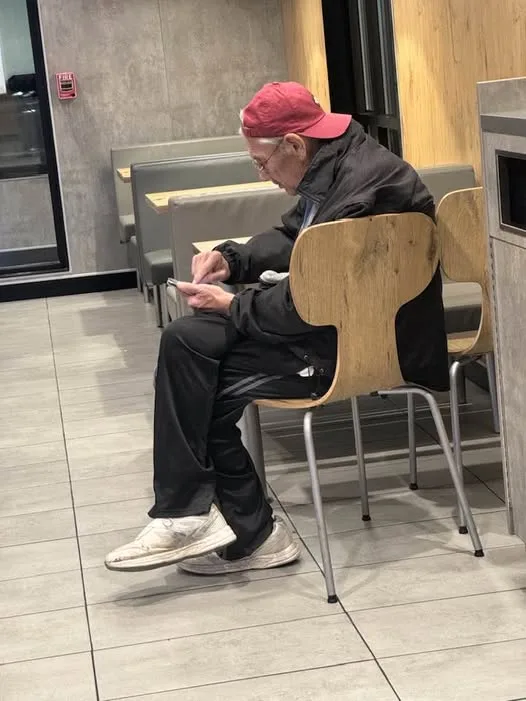 An elderly man wearing a red cap, glasses, and dark clothing sits in a modern restaurant booth, intently focused on using a…