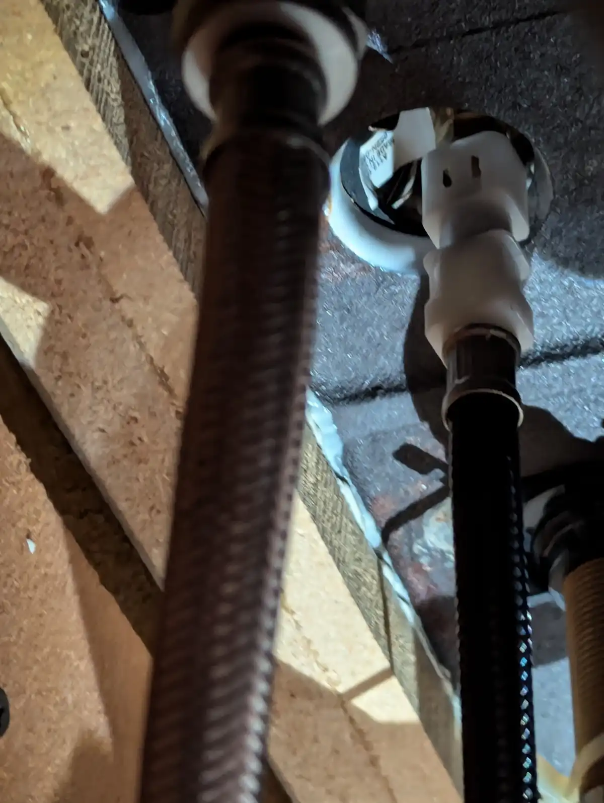 A close-up photo shows two braided metal hoses connected to a white plastic plumbing fitting under a sink, surrounded by…