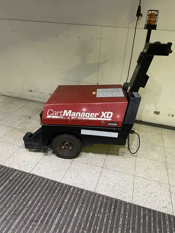 A red and black 'CartManager XD' automated cart management machine sits on a speckled tile floor next to a metal grate, with…