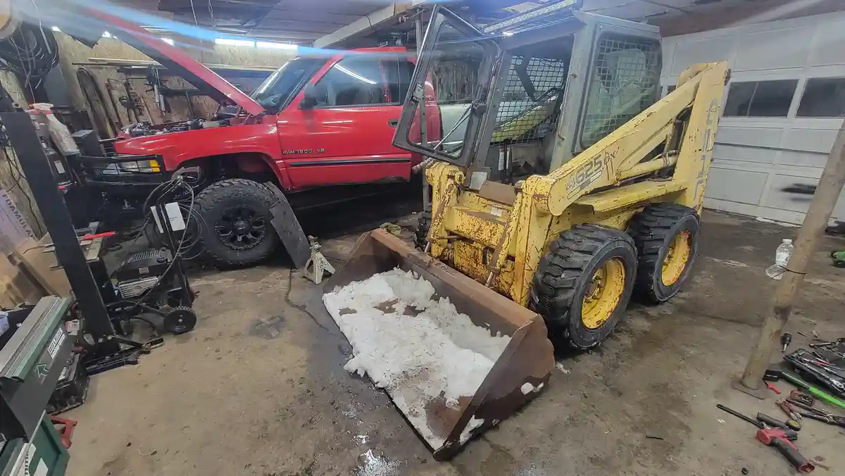A yellow skid-steer loader with a rusty bucket full of snow sits in a cluttered garage next to a red Ram 1500 pickup truck…