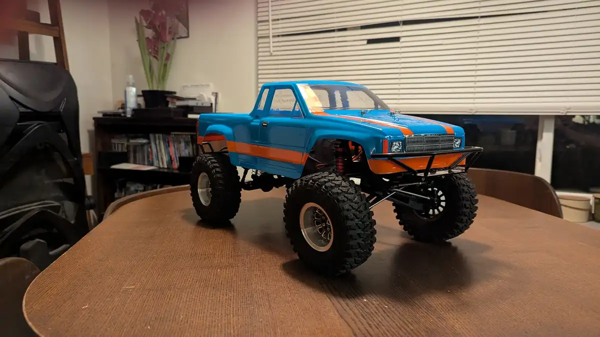 A blue and orange remote-controlled monster truck with large knobby tires sits on a wooden table in a room. The truck has a…