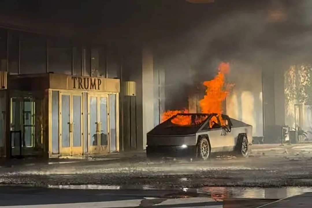 A silver Tesla Cybertruck is engulfed in flames and smoke outside a building with a prominent 'TRUMP' sign above its…