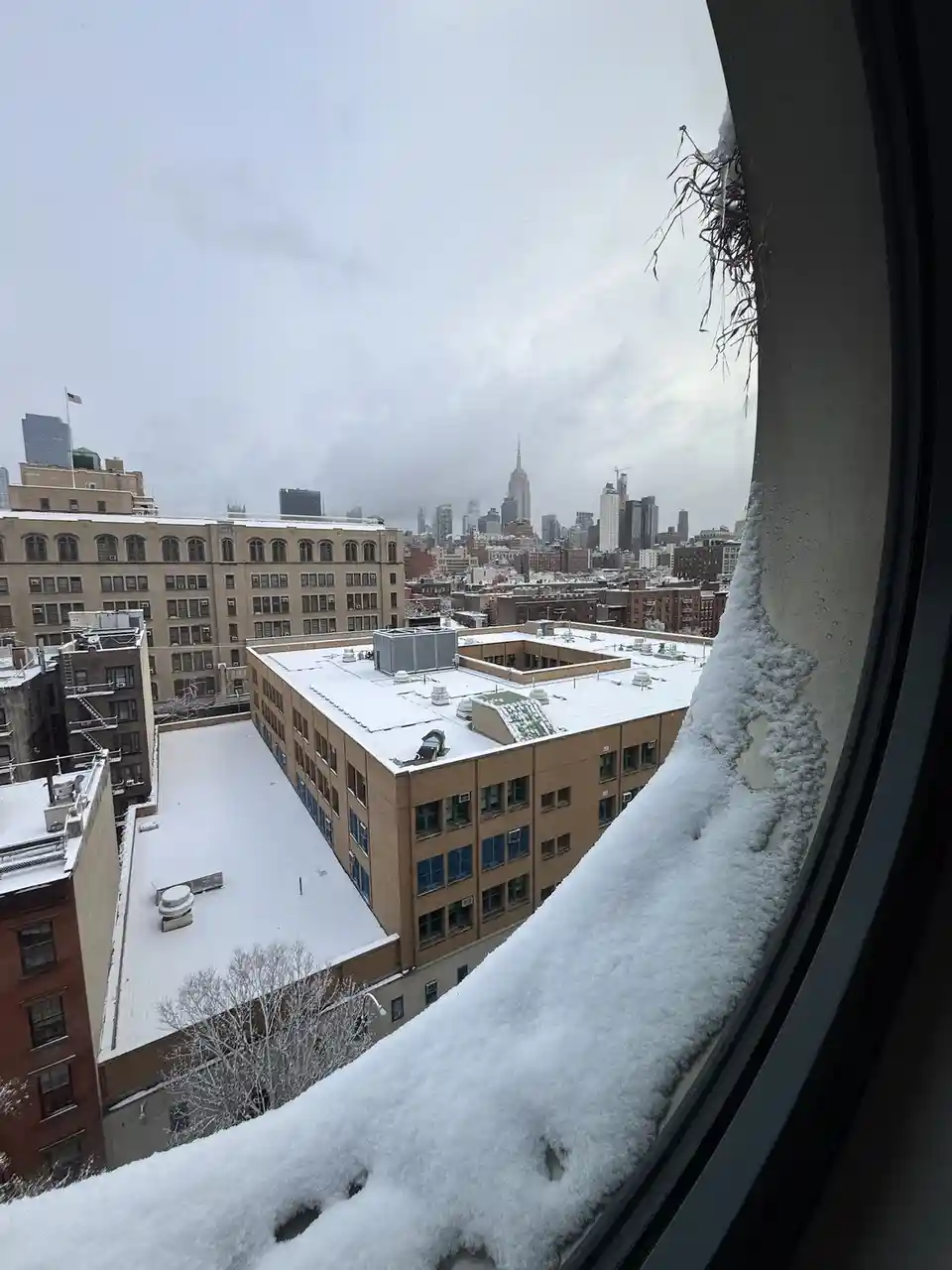 A snowy urban view from a window, with thick snow piled along the window sill and frame, overlooking snow-covered rooftops…