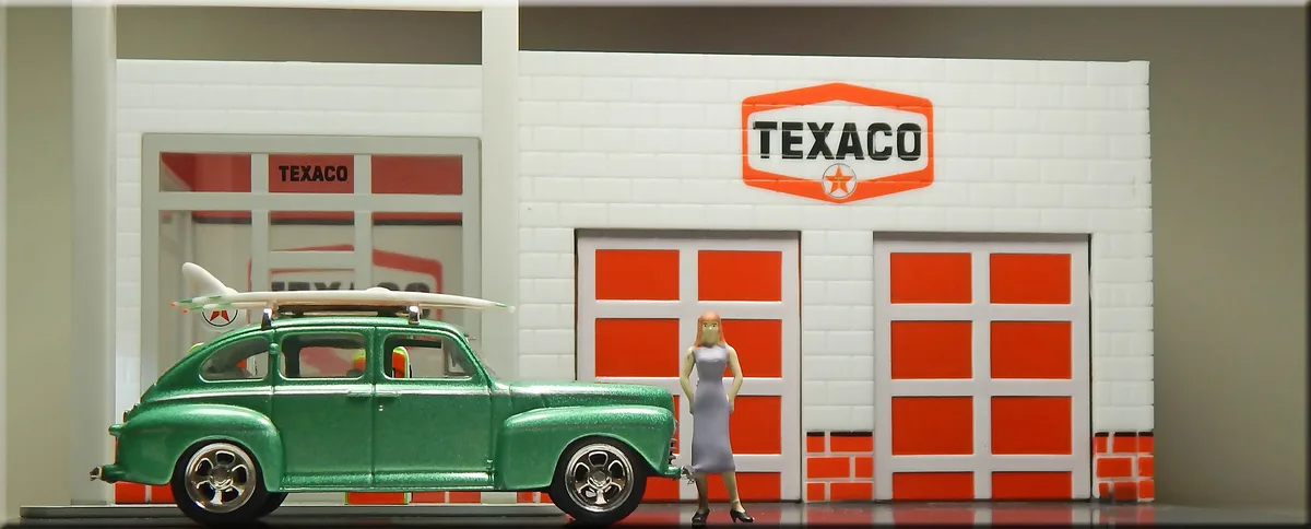 A miniature green vintage car with a surfboard on its roof is parked outside a Texaco gas station model. A miniature woman…