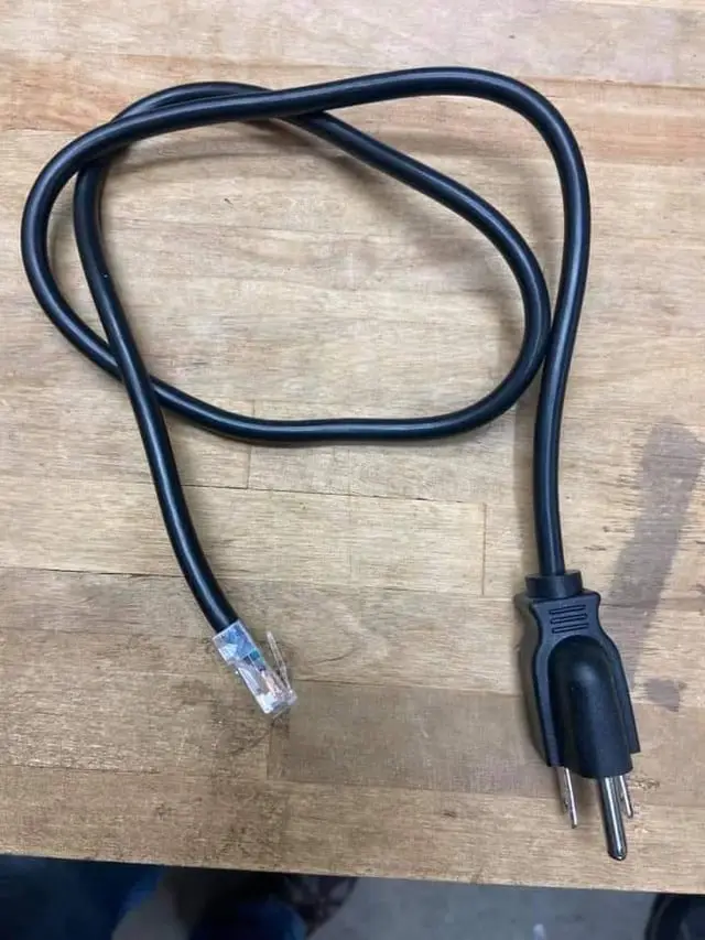 A black electrical cord lies coiled on a wooden surface, one end terminating in a standard two-prong plug and the other in…
