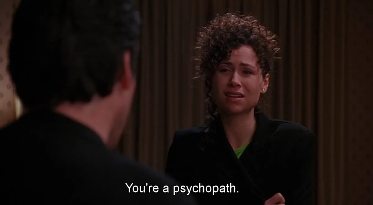 A woman with curly hair, wearing a black blazer over a green shirt, is visibly crying while facing a man whose back is to…