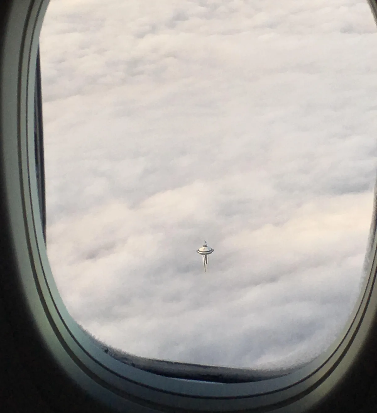 An airplane window view shows the Space Needle in Seattle emerging from a sea of clouds, appearing tiny and isolated. The…