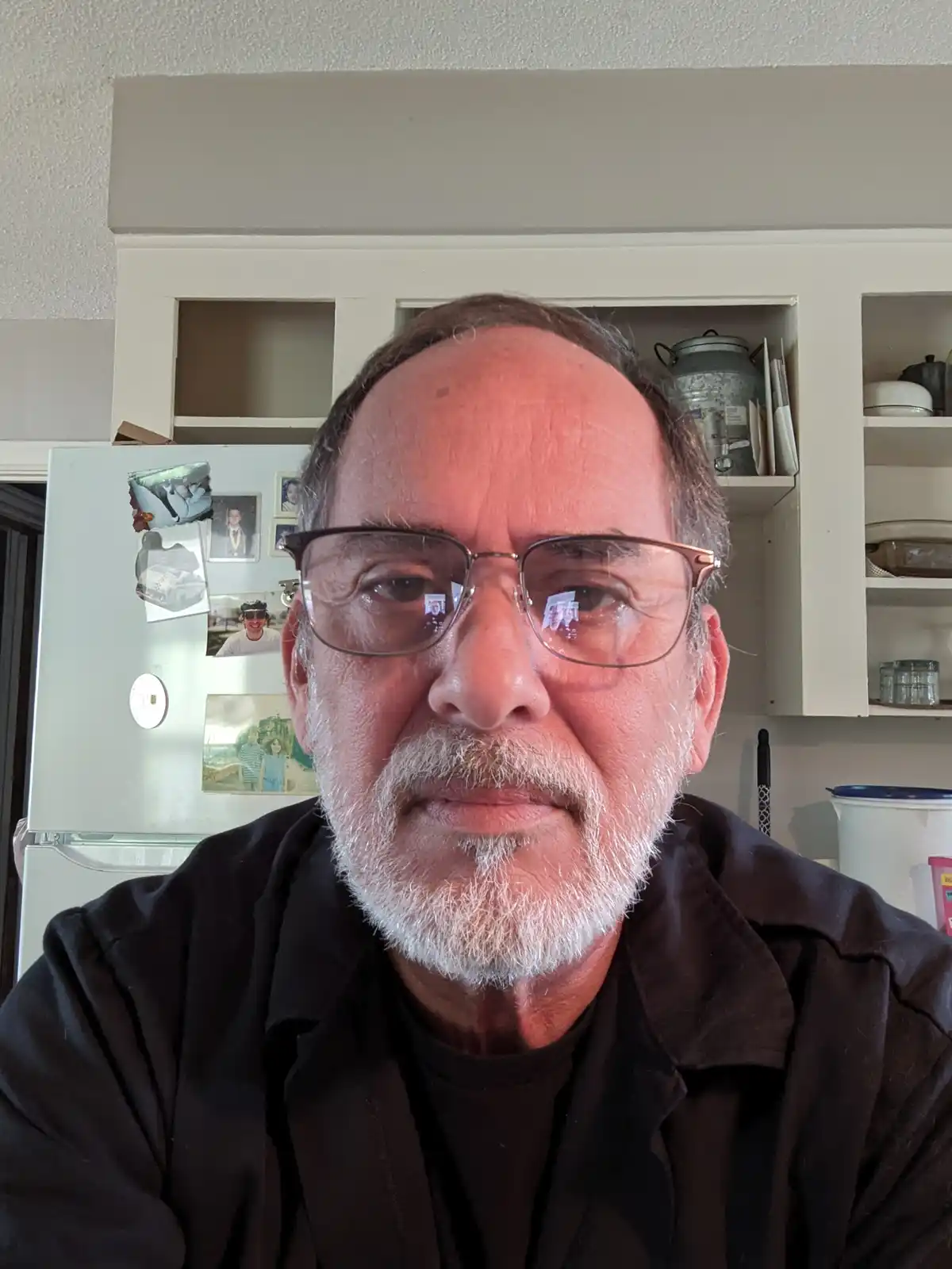A close-up selfie of a middle-aged to older man with glasses and a white beard, looking directly at the camera with a…