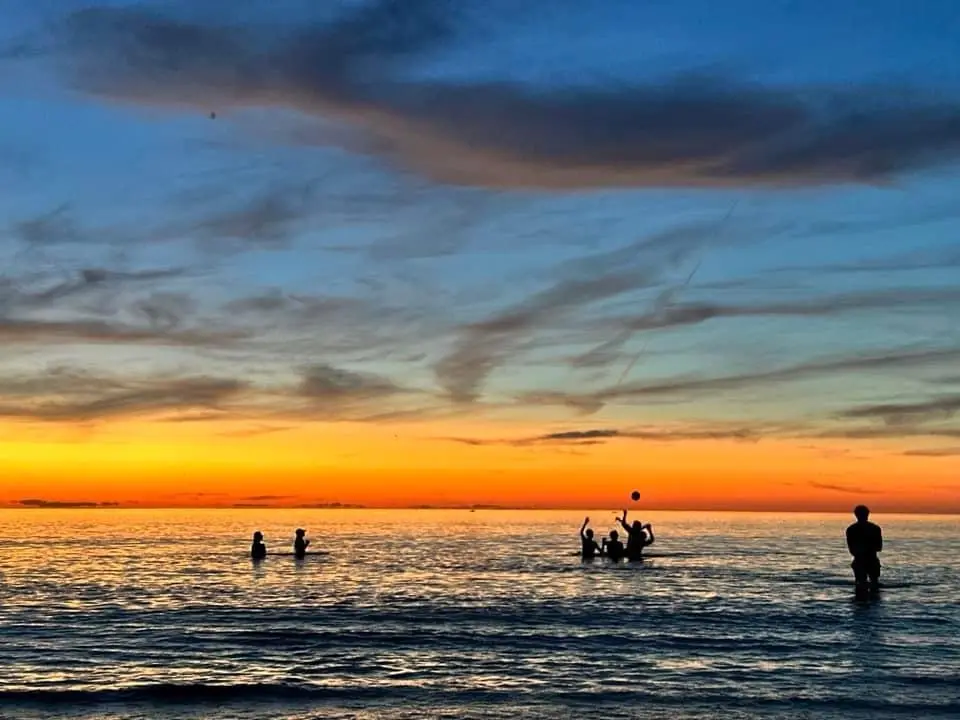 A serene sunset over the ocean with silhouettes of people playing volleyball in the water. The sky transitions from orange…