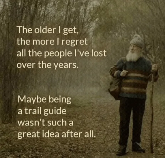 An older man with a long white beard, wearing a striped sweater and beanie, walks through a forest holding a walking stick…